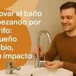 Hombre sonriente instala un grifo moderno en un lavabo, junto a un grifo viejo que será sustituido. A la izquierda, sobre un fondo con formas curvas en tonos cálidos, se muestra el título: “Renovar el baño empezando por el grifo: pequeño cambio, gran impacto”. La escena comunica renovación y mejora estética en el baño con un cambio sencillo.